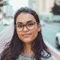 A confident woman wearing glasses smiles subtly while standing on an urban street with blurred background.